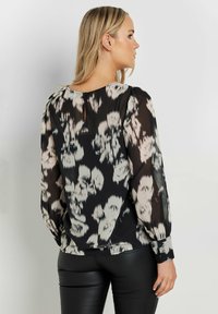 Blusa nera con motivo floreale in tonalità di crema e grigio, dotata di maniche lunghe, scollo tondo e chiusura con bottone sul retro.