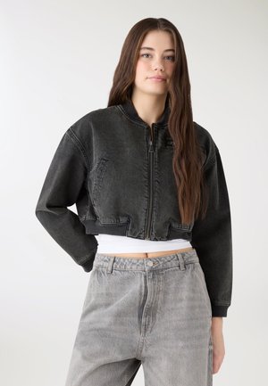 Giovane donna con capelli castani lunghi che indossa una giacca di jeans nera corta sopra una maglietta bianca e jeans grigi a vita alta, in piedi su uno sfondo semplice.