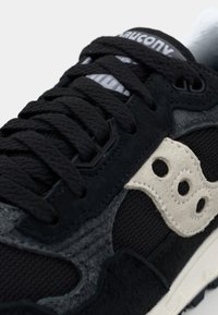 Black athletic shoe with grey and cream suede accents. Features textured laces, mesh fabric, and three-circled logo on the side.