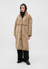 Mākslīgās kažokādas mētelis gaiši brūnā krāsā ar izteiktu faktūru, melnu apdari un lielām pogām. Tam ir augsta apkakle un garas piedurknes, to valkā virs melnas apģērba.