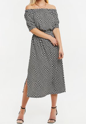Czarna i biała sukienka odsłaniająca ramiona w kropki, z krótkimi bufkami, elastyczną talią i bocznym rozcięciem, noszona z czarnymi sandałami na paski.