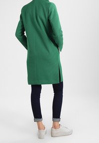 Eine Person, die einen langen grünen Mantel und dunkle Jeans trägt, steht mit dem Rücken zum Betrachter und zeigt einen lässigen Schuhstil.