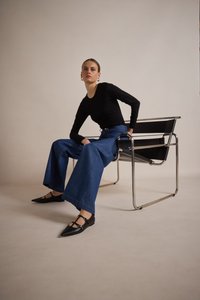 Top negro de manga larga, vaqueros de denim azules anchos con dobladillo sin acabado, bailarinas negras, sentado en una silla minimalista negra y cromo frente a un fondo neutro.
