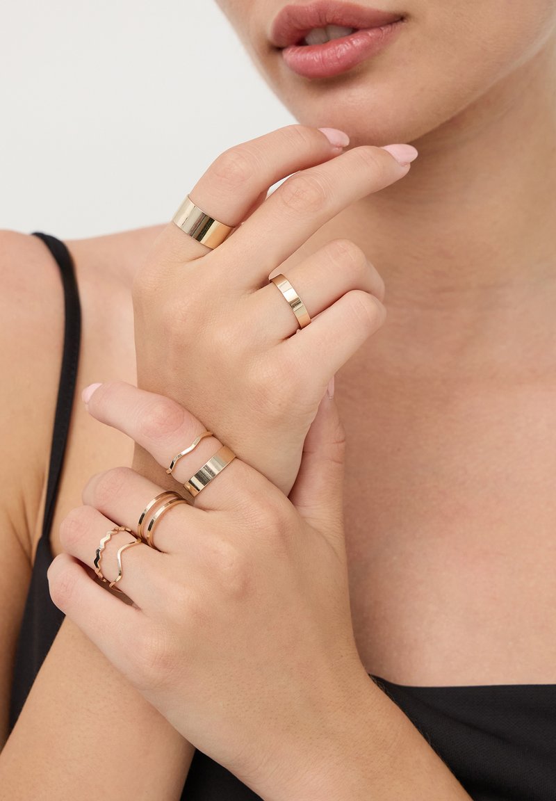 Anelli d'oro in vari design: un'ampia fascia, diverse fasce sottili e un anello con motivo ondulato. Texture lisce, superfici lucide e forme semplici.