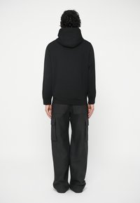 Osoba s kovrčavom kosom koja nosi crnu duksericu i crne cargo hlače, stoji okrenuta leđima prema jednostavnoj svijetloj pozadini.