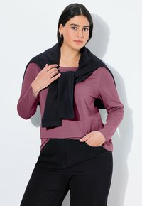 Langärmliges, mauvefarbiges Oberteil, kombiniert mit schwarzen Hosen; ein schwarzer Pullover ist lässig über die Schultern gebunden. Glatte Textur und entspannter Schnitt.