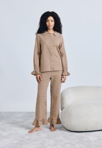 Σετ πιτζάμες καρό σε καφέ και κρεμ χρώμα με βολάν στα μανίκια και το γιακά. Χαλαρή μπλούζα και μπουφάν με φαρδιά παντελόνια, εμφανίζονται σε ανοιχτό φόντο.