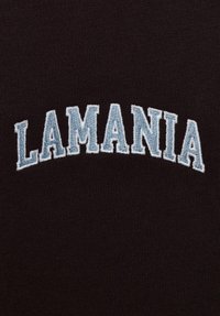 Czarna tkanina o fakturowanym, dzianinowym tle z wyhaftowanym słowem „LAMANIA” w jasnoniebieskim kolorze, łukowato przebiegającym przez środek.