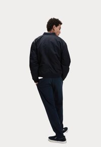 Schwarze Bomberjacke mit geripptem Kragen, seitlichen Taschen und enganliegenden Bündchen, getragen über dunklen Hosen und schwarzen Schuhen. Rückansicht dargestellt.