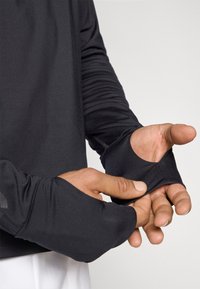 Μαύρη αθλητική μπλούζα με μακριά μανίκια, που διαθέτει ανοίγματα για τους αντίχειρες στις μανσέτες, λεία υφή και εφαρμοστό σχέδιο που αγκαλιάζει τους πήχεις.