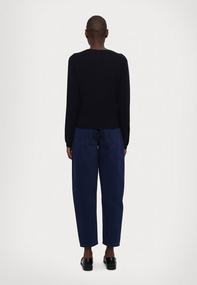 Svart strikket genser med lange ermer, rund halsåpning, tettsittende rundt livet. Kombinert med løse, mørkeblå jeans og svarte sko.