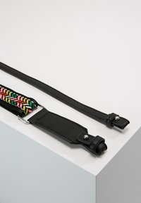 Dwa czarne skórzane paski od torebek leżą na białej powierzchni, z czego jeden ma kolorową, plecioną sekcję oraz metalową klamrę.
