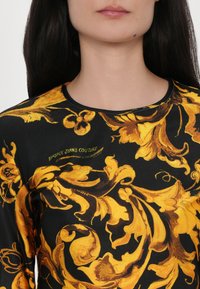 Maglietta a maniche lunghe realizzata in tessuto nero decorato con luminosi motivi dorati ornate, colletto rotondo e un discreto dettaglio del logo nel design.