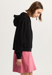 Svart cropped hoodie med dragsko, kombinerat med en rosa knälång kjol. Tyget verkar mjukt, med en slät yta och enkelt mönster.