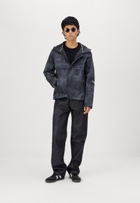 Zwarte gestreepte jas met capuchon, ritsopening aan de voorkant en zijzakken, gecombineerd met donkere jeans en zwarte sneakers. Ronde zonnebril gedragen.