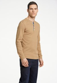 Långärmad, beige henley skjorta med tre vita knappar och en blå accent vid halsringningen, i kombination med mörka denimjeans.