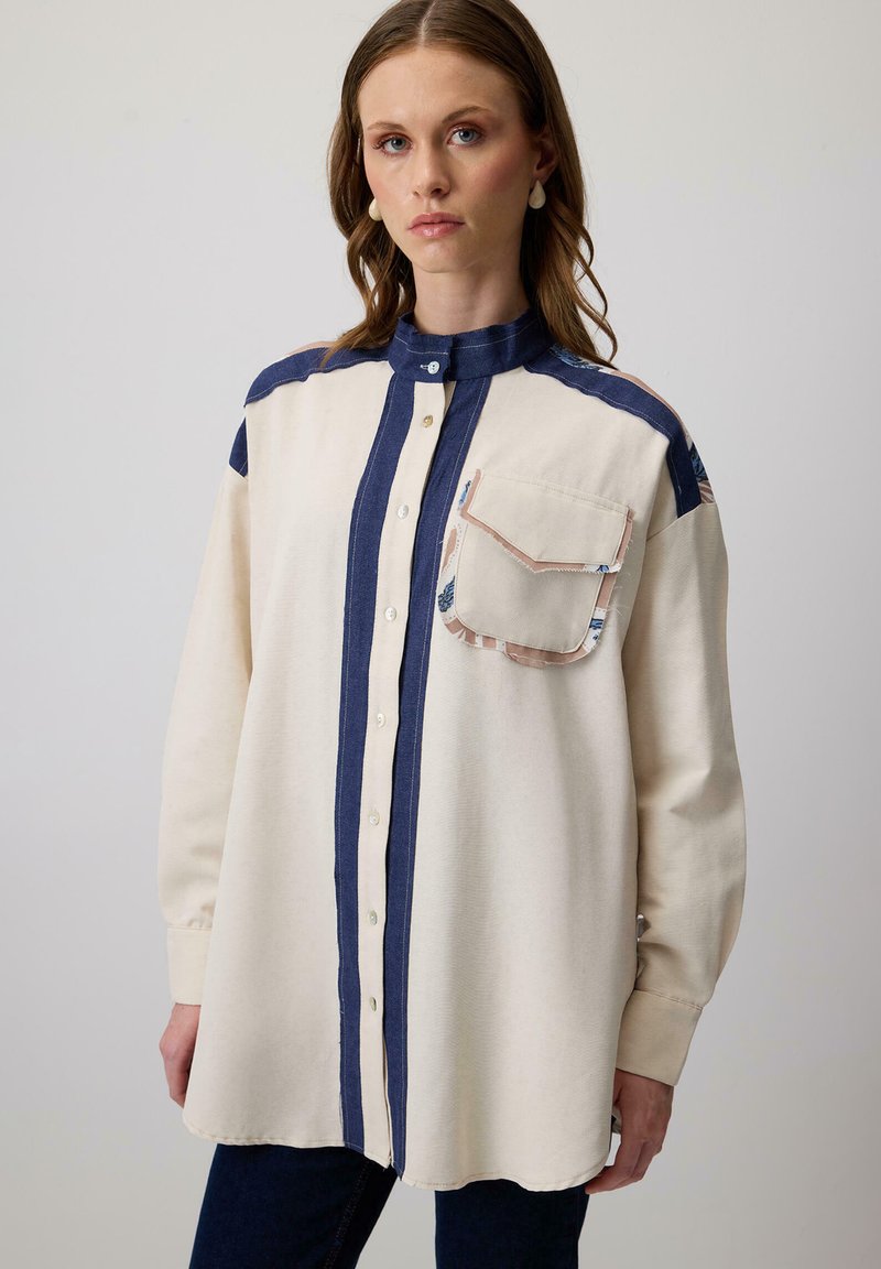 Crème en marine knoopblouse met hoge kraag, voorzien van een borstzak, contrasterend paneelontwerp en een zachte stoftextuur.