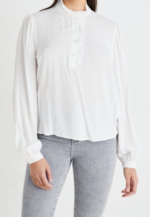 Kvinde iført en hvid langærmet bluse med teksturerede prikker og flæset krave kombineret med lysegrå tætsiddende jeans.