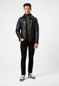 Schwarze Lederjacke mit Reißverschluss, grüner Rollkragenpullover, schwarze schmal geschnittene Hose und schwarz-weiße Sneakers mit Schnürung.