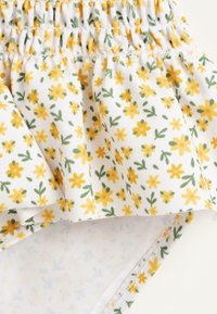 Tissu blanc avec un motif floral jaune. Présente un élastique froncé en haut et une texture douce. Des feuilles vertes supplémentaires rehaussent le design.