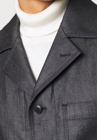 Chaqueta de denim gris oscuro con cuello de muesca, cierre de botones en el frente y un bolsillo en el pecho, superpuesta sobre un suéter de cuello alto blanco.