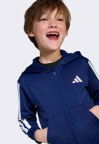 Námořnická zipová mikina s bílými pruhy na rukávech, s přední kapsou a kapucí. Vyrobena z hladkého materiálu, s logem na hrudi.