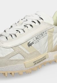 Λευκό αθλητικό παπούτσι Lacoste με μπεζ λεπτομέρειες, μεταλλικά κορδόνια, λογότυπο κροκόδειλου και κείμενο με συντεταγμένες στην υφασμένη σόλα.
