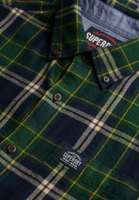 Grün-karierte Flanellhemd in Marineblau, Gelb und Weiß mit braunen Knöpfen und einem Superdry Work-Wear-Label auf der Brusttasche.