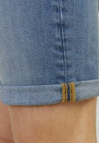 Pantalones cortos de mezclilla de color azul claro, con dobladillos enrollados y acentos de costura en marrón. La tela tiene una textura con un tejido de mezclilla clásico.