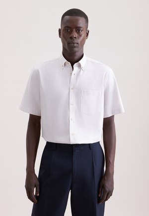 Mannelijk model met een wit, kort overhemd met knopen en een borstzak, gecombineerd met donkerblauwe broek, staand tegen een effen achtergrond.