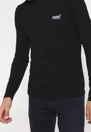 Mężczyzna ubrany w dopasowaną czarną koszulę z długim rękawem z małym białym logo "Super Dry" na klatce piersiowej oraz ciemne spodnie.