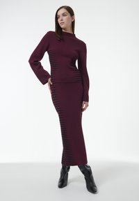 El conjunto de vestido y top de punto burdeos presenta un cuello alto, mangas largas y detalles en zigzag negros a lo largo del costado. Combinado con botas de tacón negras.