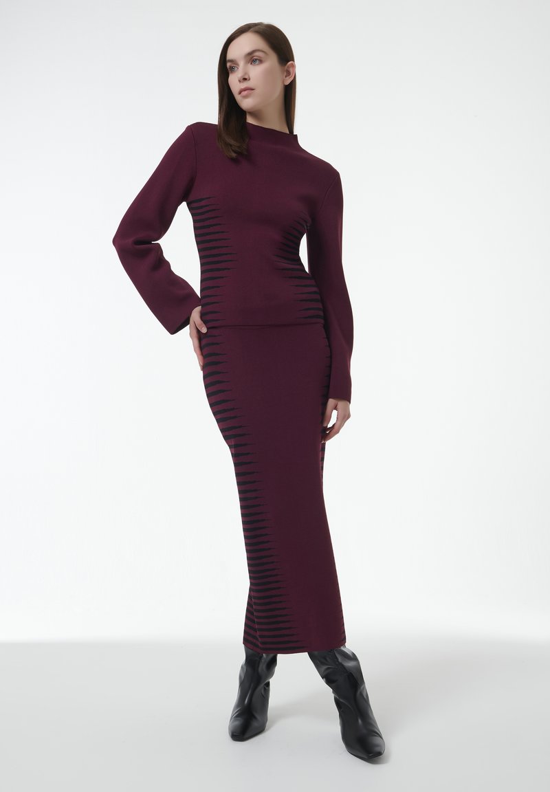 El conjunto de vestido y top de punto burdeos presenta un cuello alto, mangas largas y detalles en zigzag negros a lo largo del costado. Combinado con botas de tacón negras.