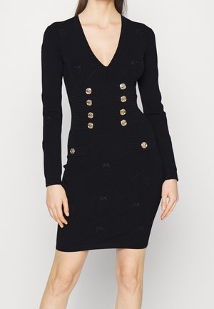 Czarna dopasowana sukienka z długimi rękawami, głębokim dekoltem w serek oraz dwoma rzędami złotych guzików z przodu i przy biodrach, noszona przez stojącą kobietę.