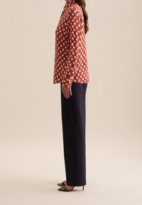 Kvinna som står på sidan, klädd i en långärmad röd och vit mönstrad blus, svarta vida byxor och svarta högklackade sandaler.