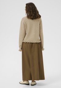 Ženska s kravljevimi lasmi do ramen, oblečena v bež pulover s volančki, rjavo dolgo krilo in smetanaste loaferje, obrnjena stran od kamere na enobarvni ozadju.