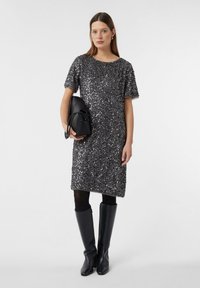 Vrouw in een grijze, glinsterende jurk met korte mouwen tot de knie, zwarte panty's, zwarte laarzen tot de knie, die een zwarte handtas vasthoudt tegen een witte achtergrond.