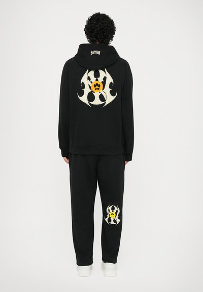 Čierna súprava s mikinou a teplákmi. Mikina má na chrbte veľkú vyšívanú bielo-žltú grafiku; tepláky majú menšiu zodpovedajúcu grafiku.