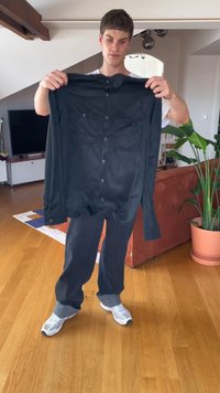 Jovem dentro de casa segurando uma camisa preta de botões à frente de um sofá, planta em vaso e televisão sobre um chão de madeira.