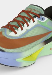 Το παπούτσι για τρέξιμο Nike Zoom Fly σε πράσινο, καφέ και κίτρινο με κόκκινα κορδόνια και λεπτομέρεια από διαπνέον ύφασμα, εμφανίζοντας τη πλευρική και μπροστινή γωνία.