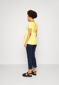 Top amarelo de mangas curtas com corpete texturizado e estilo peplum solto. Usado com jeans azul escuro e sandálias pretas, visto de lado.