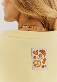 Žltá mikina s textúrovanou tkaninou, na ktorej je biely pruh s hnedými ilustráciami jedla, vrátane srdca, praclíka a pečiva.