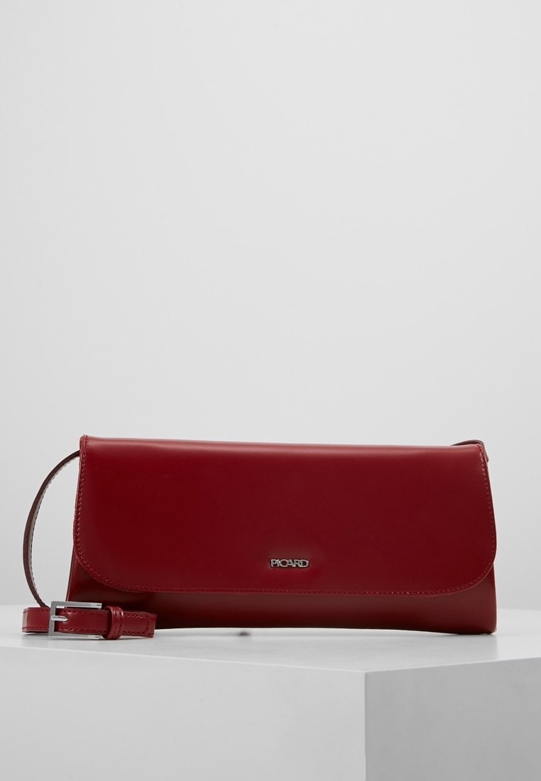 Pochette rectangulaire en cuir rouge avec une sangle à boucle argentée et un petit logo argenté sur le devant, présentée sur une surface blanche.