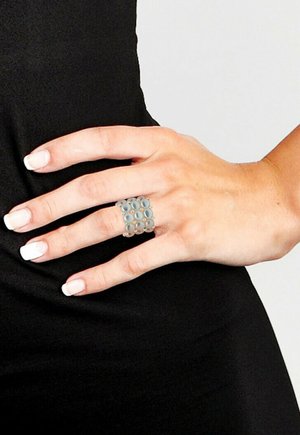 Main avec manucure française portant une large bague en argent avec des motifs circulaires, reposant sur un fond en tissu noir.