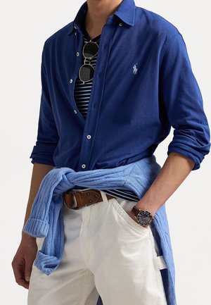 Mężczyzna w granatowej koszuli na guziki noszonej na pasku w paski, jasnoniebieski sweter zawiązany w pasie, beżowe spodnie, brązowy pasek, zegarek i okulary przeciwsłoneczne zawieszone na koszuli.