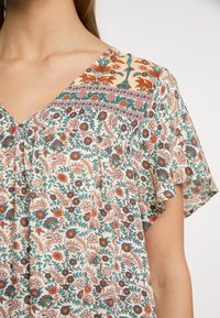 Bluse mit floralen Druck in Creme, mit V-Ausschnitt und kurzen Rüschensäumen. Die Farben umfassen Teal, Orange und Pink mit detaillierten Mustern.