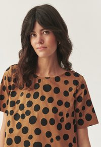 Una donna con capelli mossi e scuri indossa una camicia marrone con vari punti neri, espressione neutra, in piedi contro uno sfondo chiaro e uniforme.