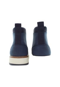 Ett par marinblå ankelskor med svarta dragflikar och beige sulor, sedda bakifrån mot en vit bakgrund.