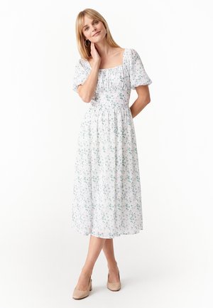 Blonde vrouw draagt een witte midi-jurk met kleine groene bloemenprints en beige hakken, poserend met één hand die haar nek raakt.