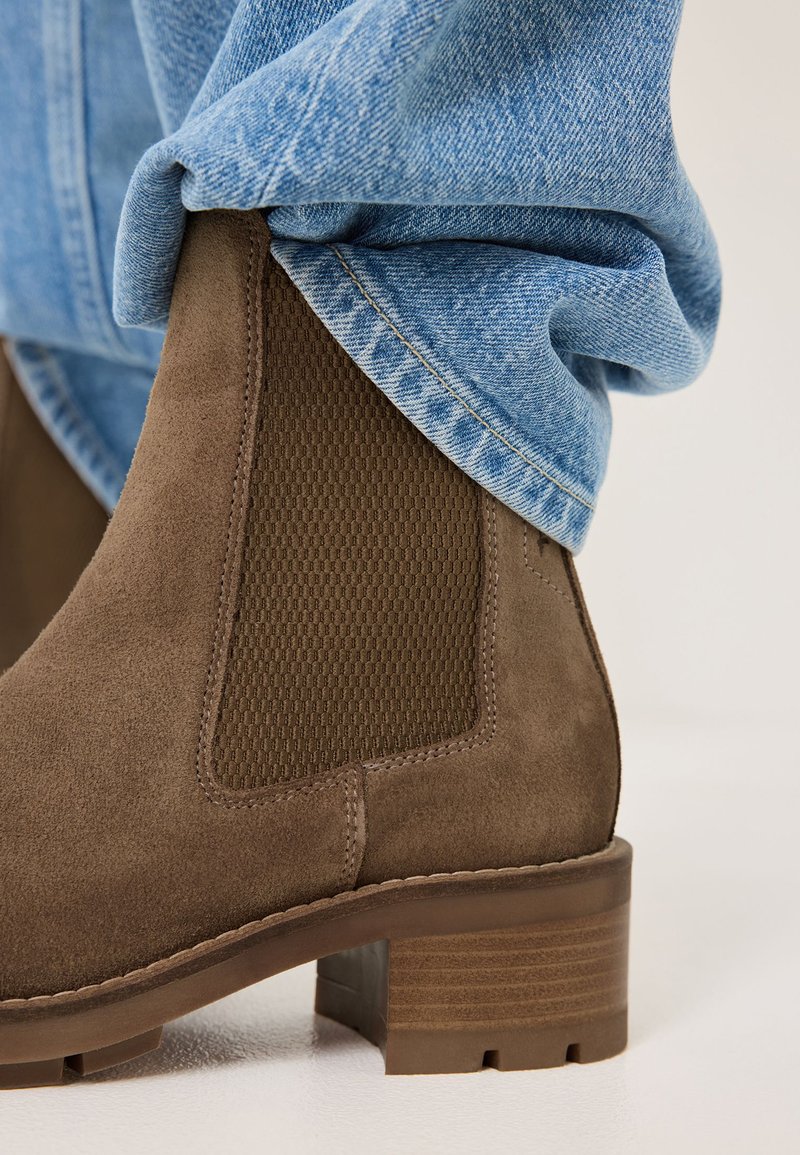 Bottine en daim marron avec un panneau latéral en élasthanne texturé et un talon bloc robuste, associée à un jean en denim bleu clair retroussé.